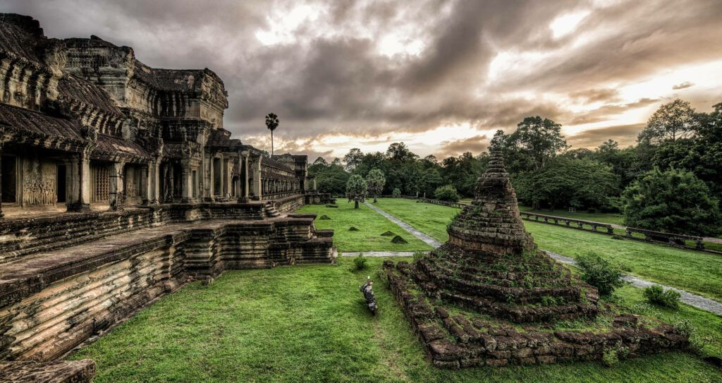Ancient Angkor Wat ruins surrounded by lush green grass and vibrant nature under cloudy sky Wallpaper