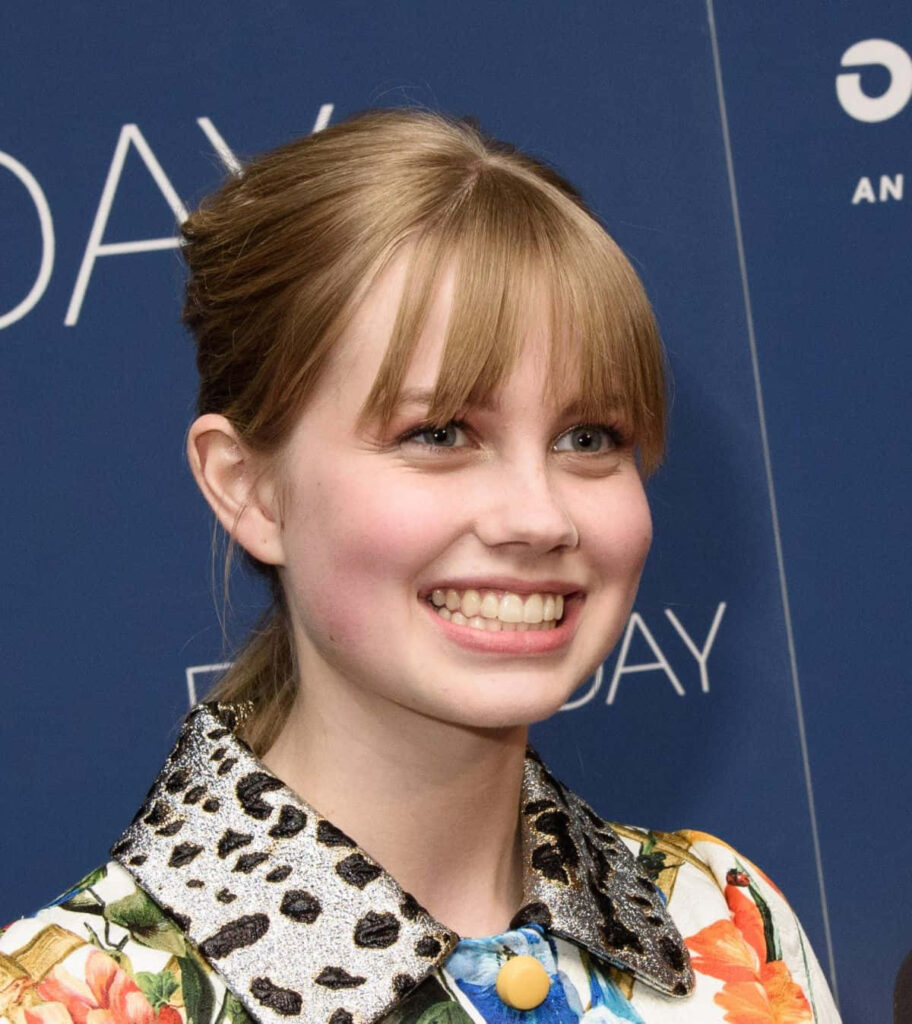 Angourie Rice smiling in colorful floral outfit at event with vibrant blue background wallpaper