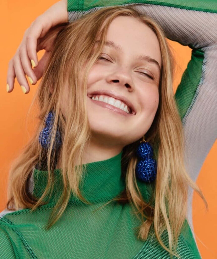 Angourie Rice smiling wearing green top and bold blue earrings on vibrant orange background wallpaper