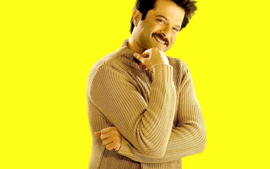 Anil Kapoor smiling brightly in light brown sweater on vibrant yellow wallpaper background