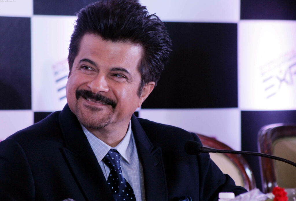 Anil Kapoor smiling confidently in formal suit at press conference with checkered backdrop wallpaper