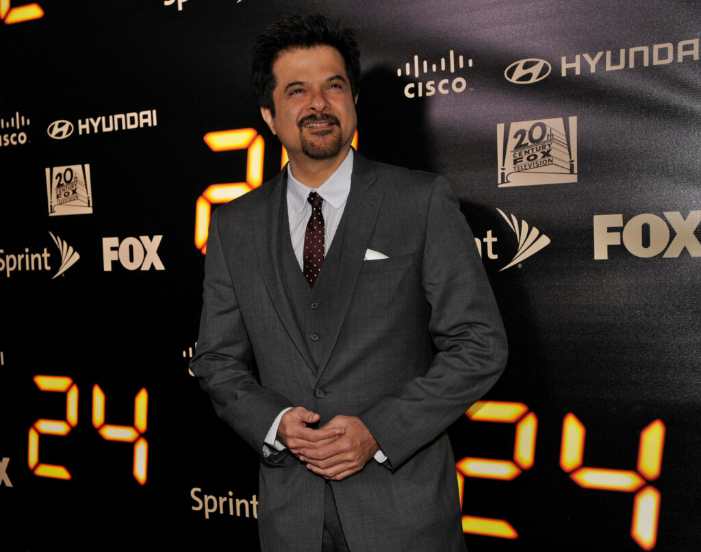 Anil Kapoor in sharp grey suit posing confidently with number 24 and network logos background Wallpaper