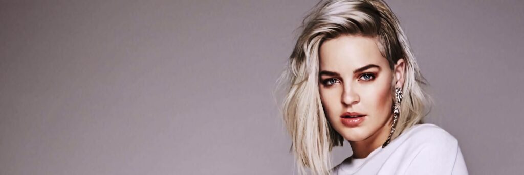 Anne Marie with platinum blonde hair posing confidently against a soft neutral wallpaper background