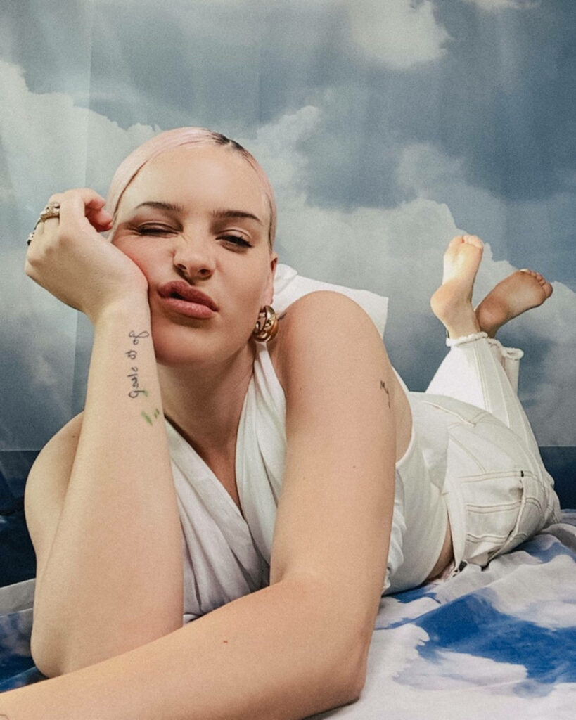 Anne Marie smiling lying on bed with cloud wallpaper background playful confident photoshoot