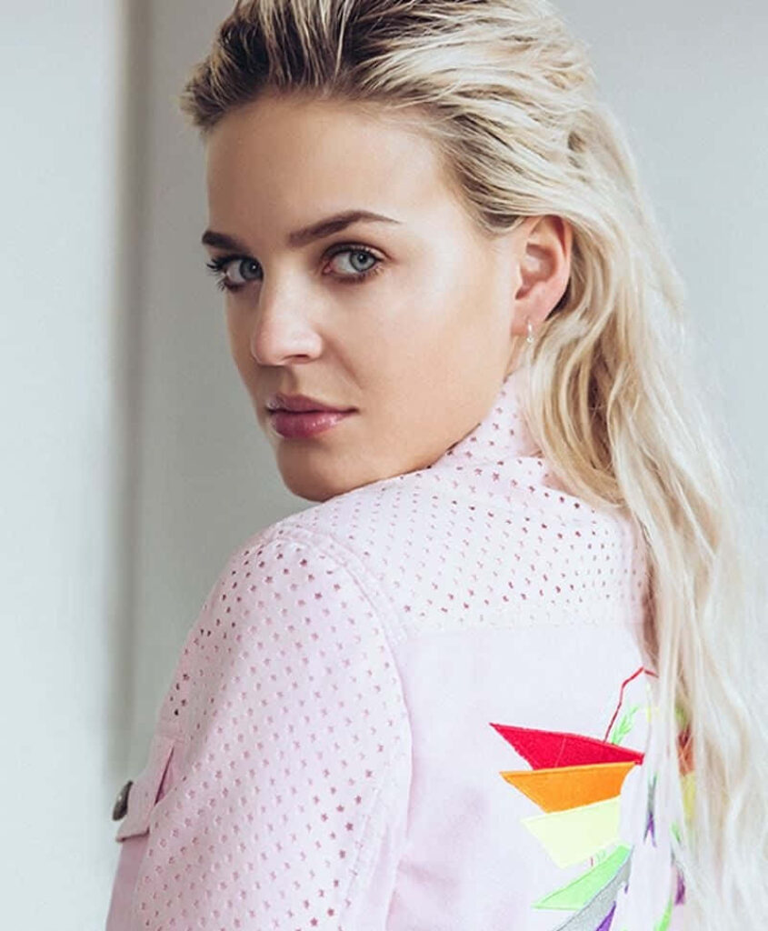 Anne Marie in light pink jacket with colorful geometric patterns vibrant modern wallpaper background