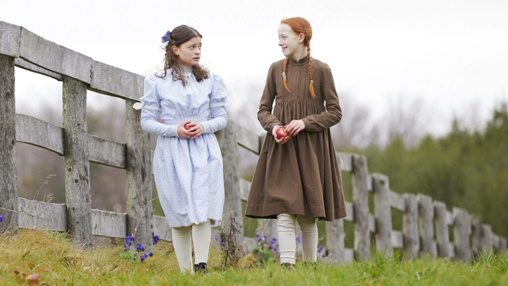 Anne in classic period attire walking in grassy field with friend warm nostalgic Anne With An E wallpaper
