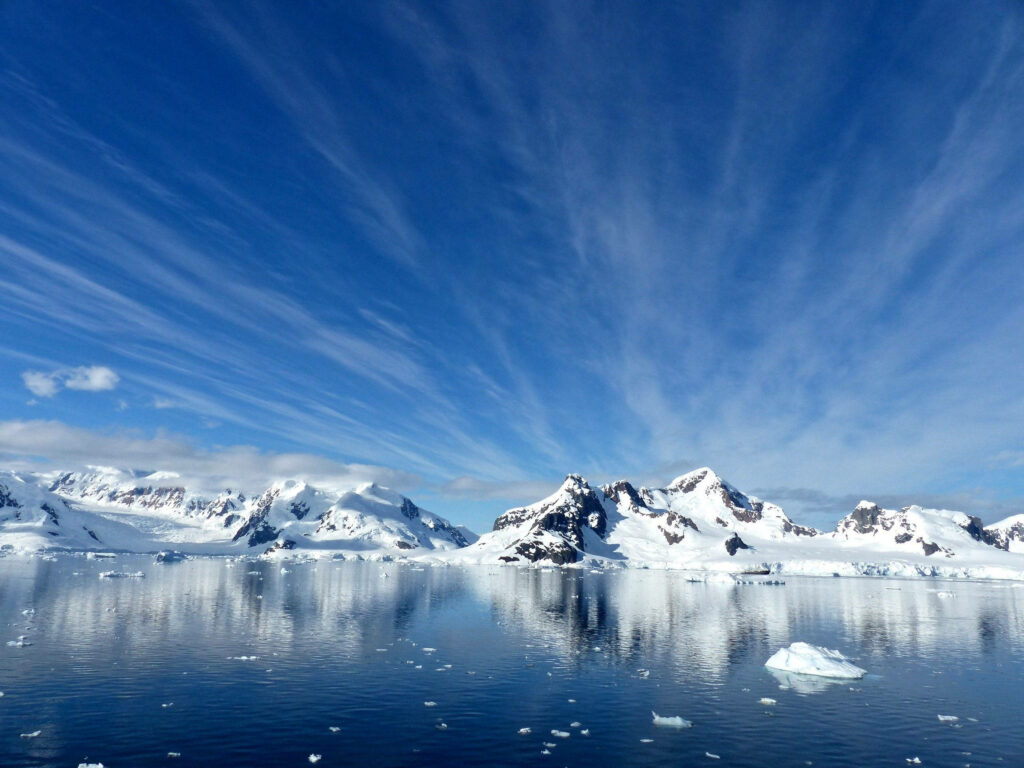 Antarctica snow-covered mountains reflected on calm icy water under bright blue sky with clouds Wallpaper
