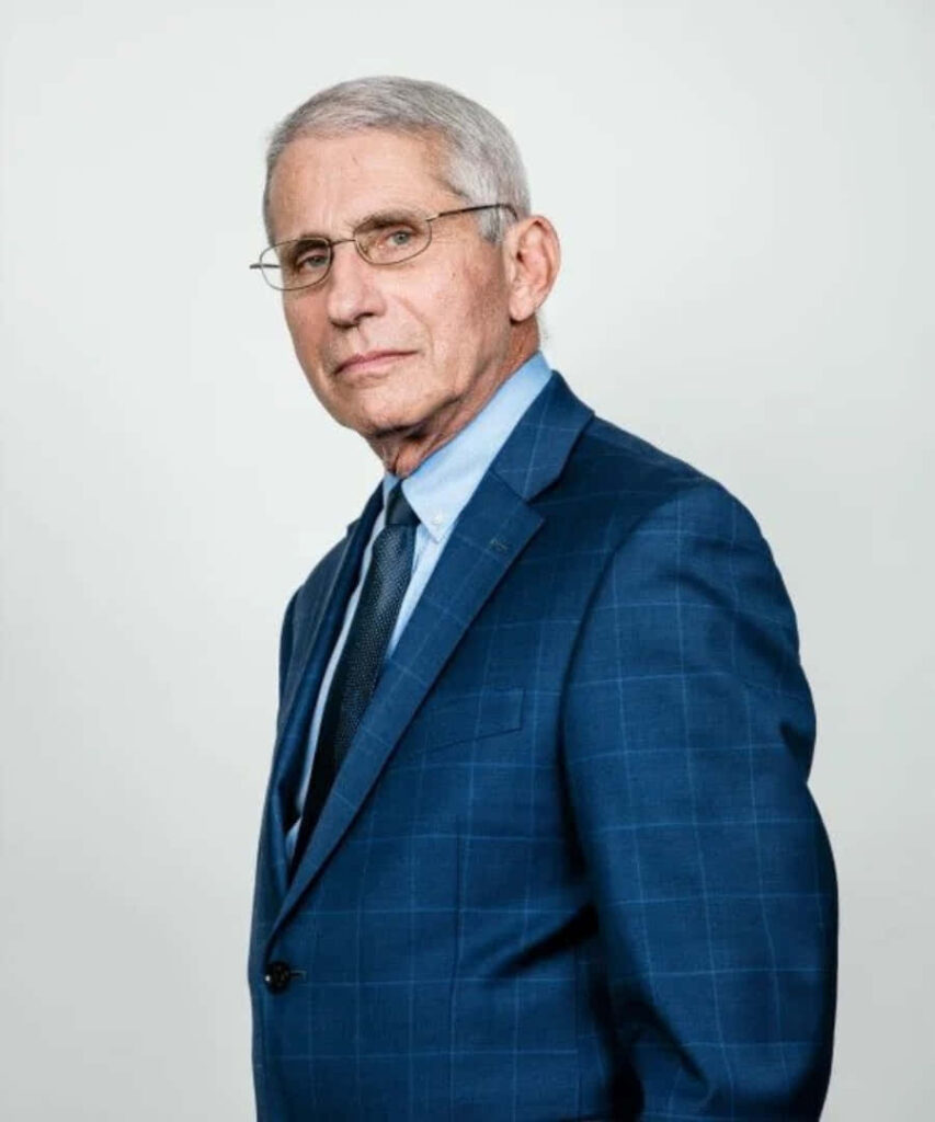 Anthony Fauci in blue suit confidently addressing media at press conference with wallpaper background