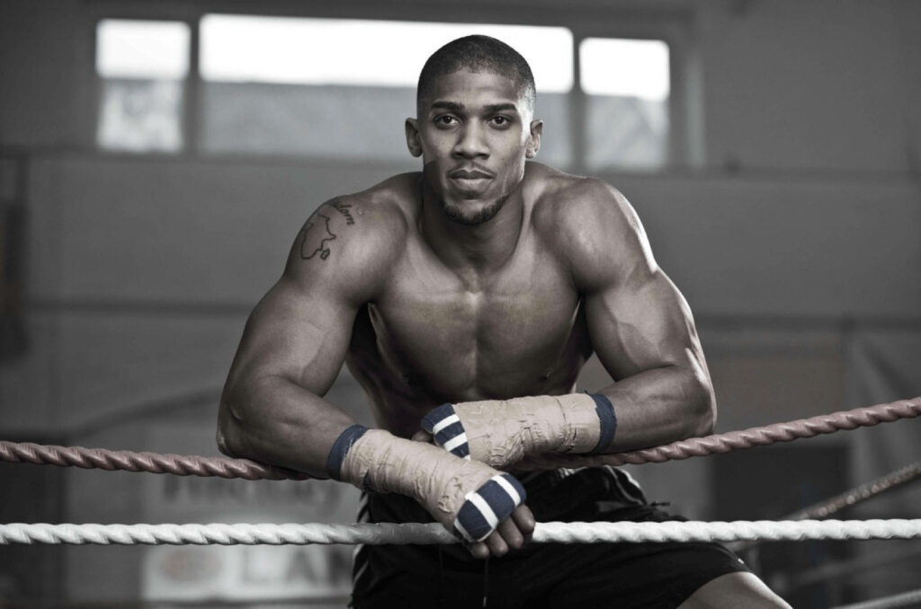 Anthony Joshua leaning on boxing ring ropes showing powerful physique and intense focus background Wallpaper
