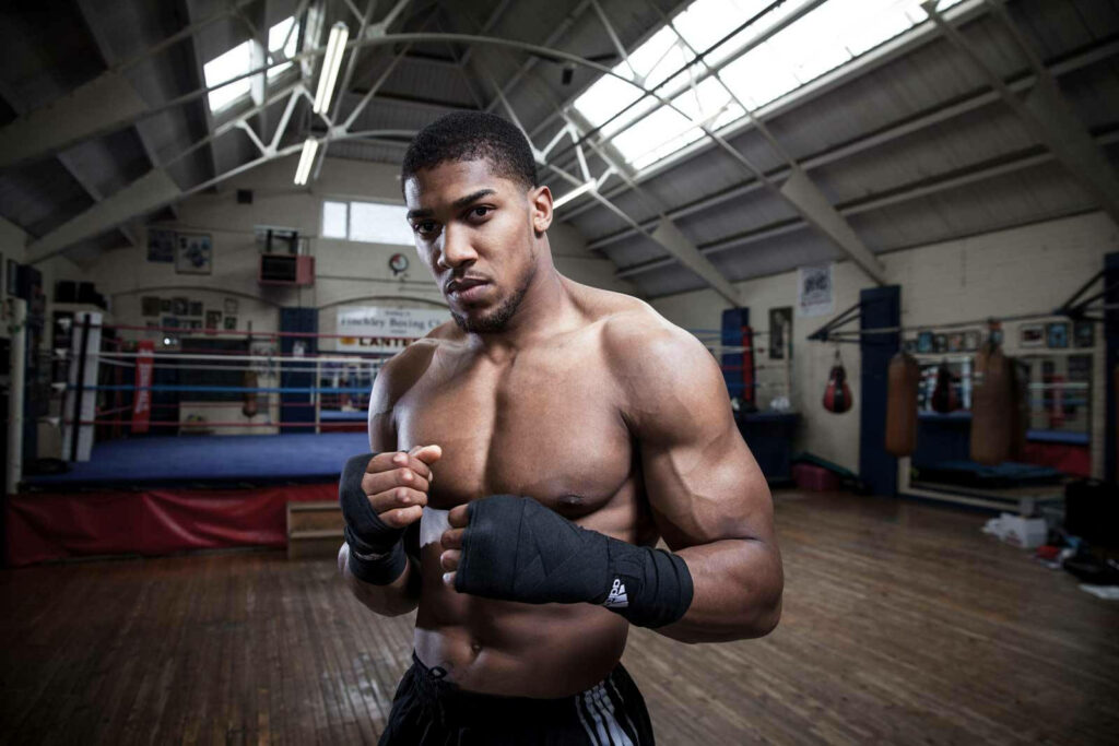 Anthony Joshua shirtless focused in gym boxing gloves wrapped with boxing ring background wallpaper