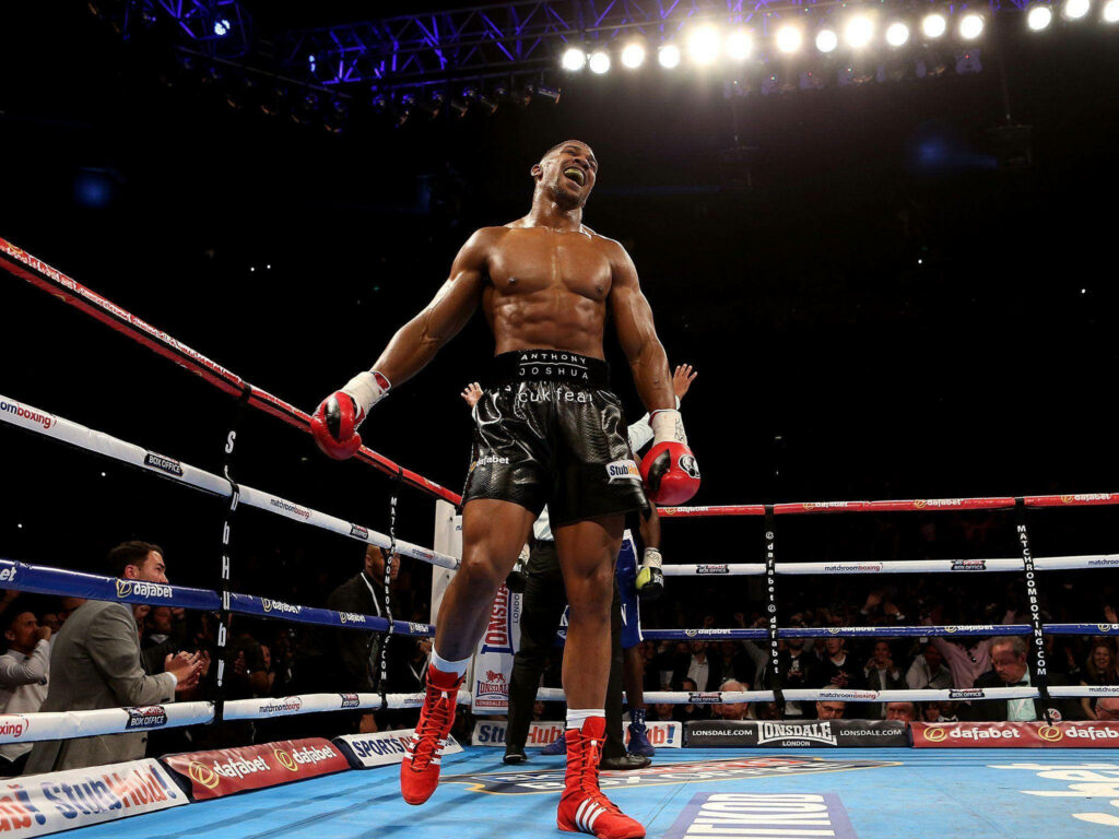 Anthony Joshua triumphant by boxing ring ropes showing strength and athleticism under lights Wallpaper