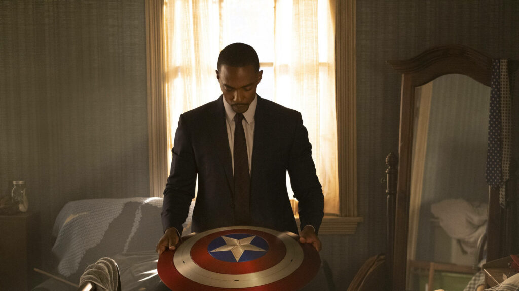 Anthony Mackie in suit holding Captain America shield in dim room reflecting legacy wallpaper