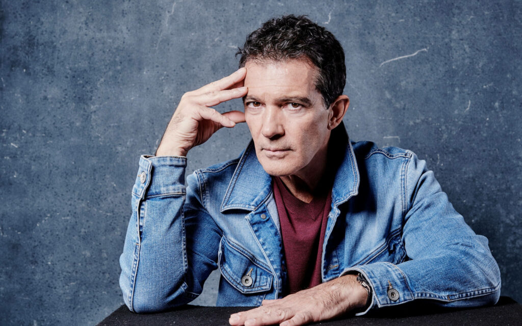 Antonio Banderas in denim jacket posing thoughtfully against textured blue wallpaper background