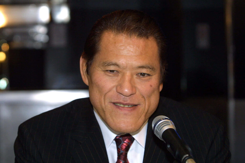 Antonio Inoki smiling confidently in sharp suit at press event with microphone and wallpaper background