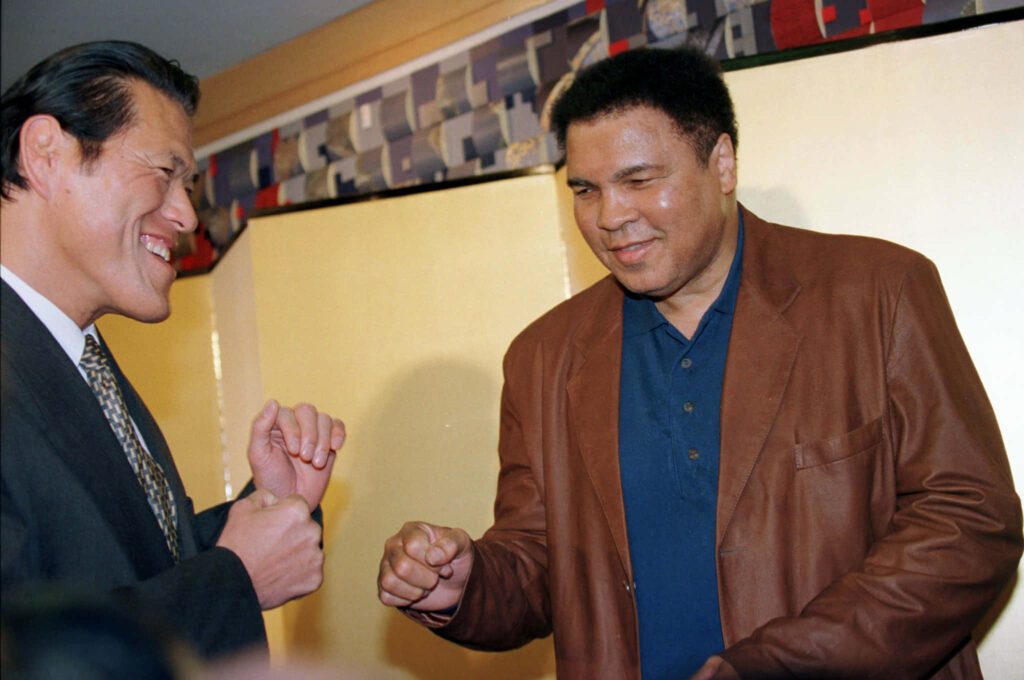 Antonio Inoki smiling and sharing a friendly moment with another person in a warm wallpaper background