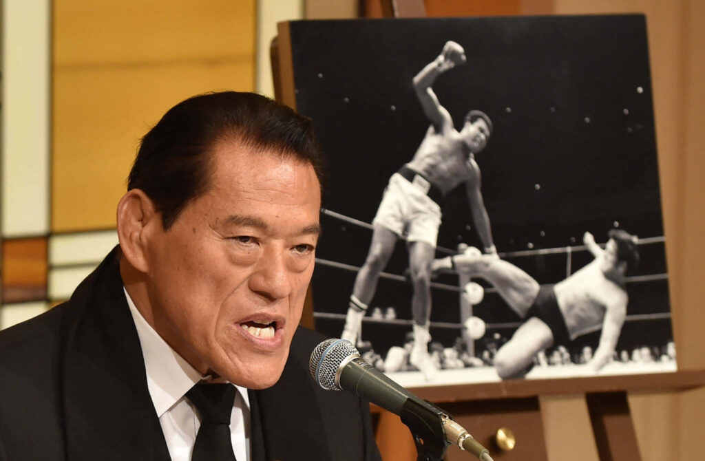 Antonio Inoki speaking at podium with iconic wrestling match photo highlighting his legacy wallpaper