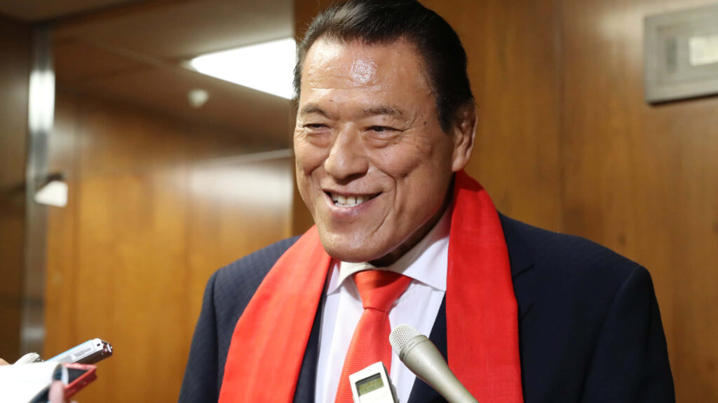 Antonio Inoki smiling at media conference wearing suit with red scarf and tie wood panel background Wallpaper