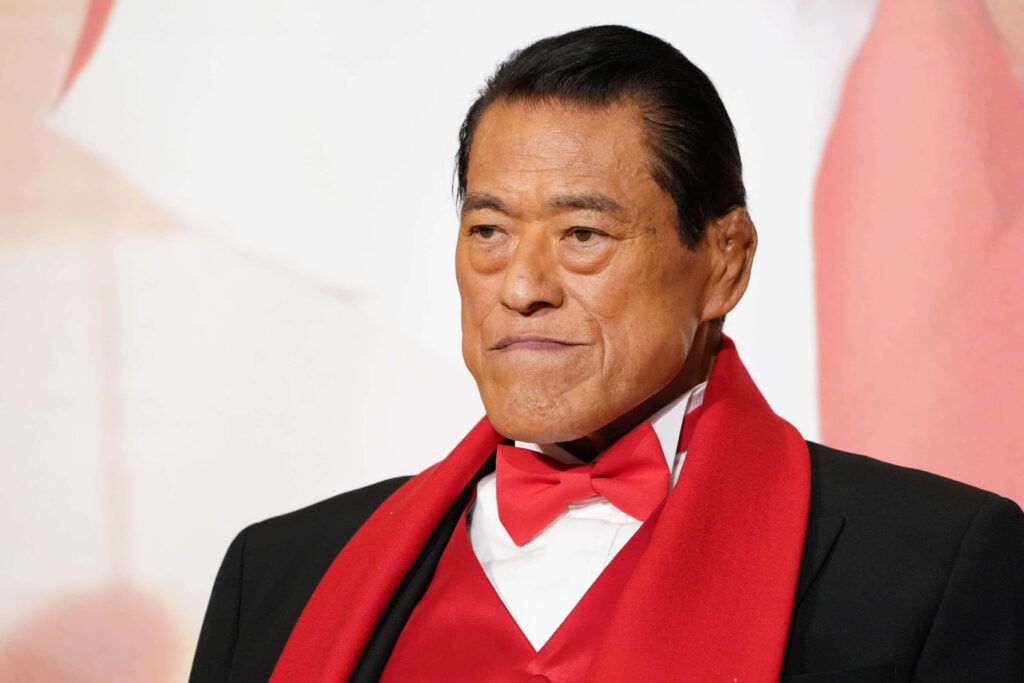 Antonio Inoki in black suit with red bow tie and scarf striking red and black wallpaper background