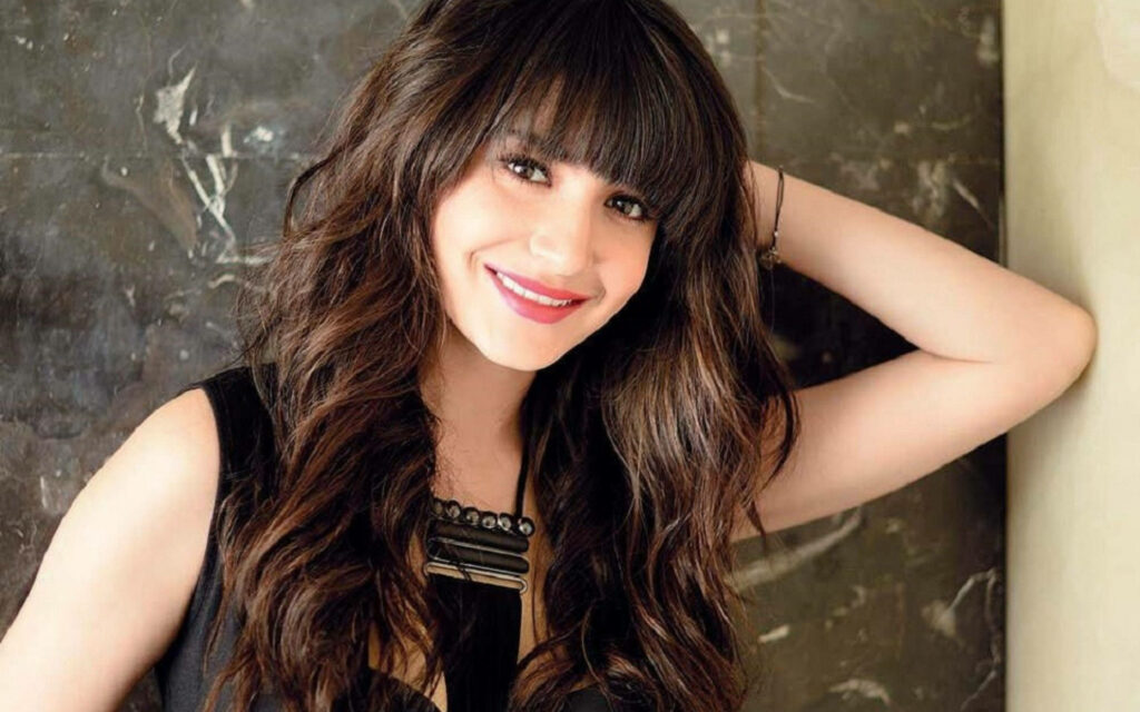 Anushka Sharma with soft curls and stylish bangs posing against textured marble wallpaper background