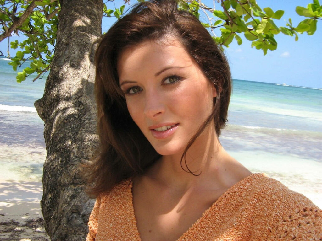 April Bowlby on sandy beach with turquoise waves under bright sky near lush tropical tree Wallpaper
