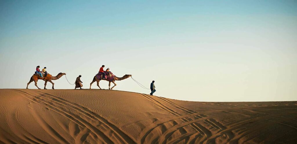 Arab man leading camel across golden desert dunes under clear blue sky panoramic wallpaper background
