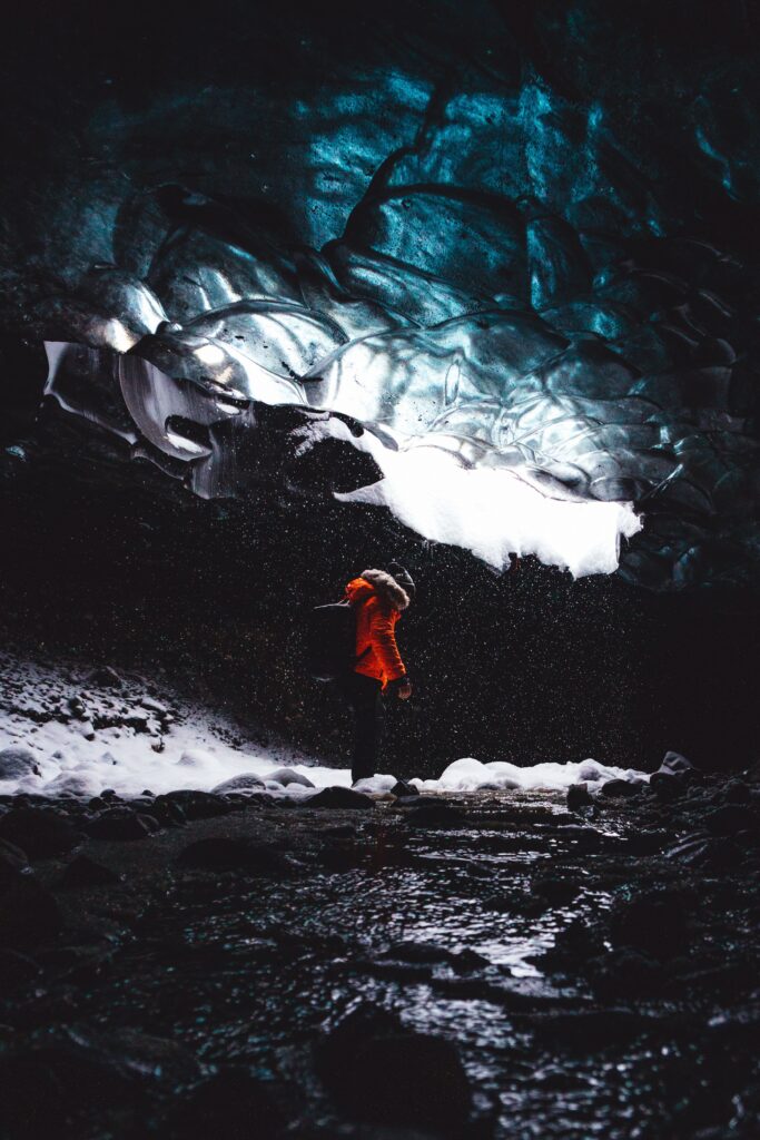 Man in bright orange jacket inside icy blue Arctic cave with snow and ice floes winter adventure Wallpaper