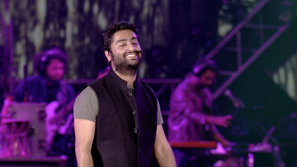 Indian singer Arijit Singh smiling on stage during lively concert with vibrant wallpaper background