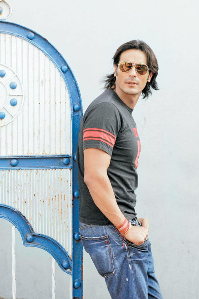 Arjun Rampal leaning on blue white gate wearing sunglasses black t shirt red stripes blue jeans Wallpaper