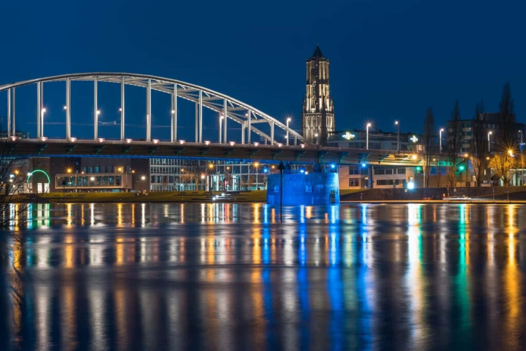 Arnhem Bridge at night glowing with vibrant lights and shimmering reflections on calm river Wallpaper