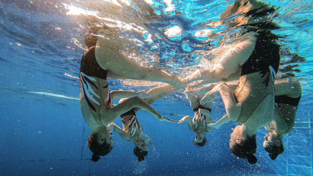 Artistic swimming team performing synchronized underwater moves with clear blue pool reflections Wallpaper