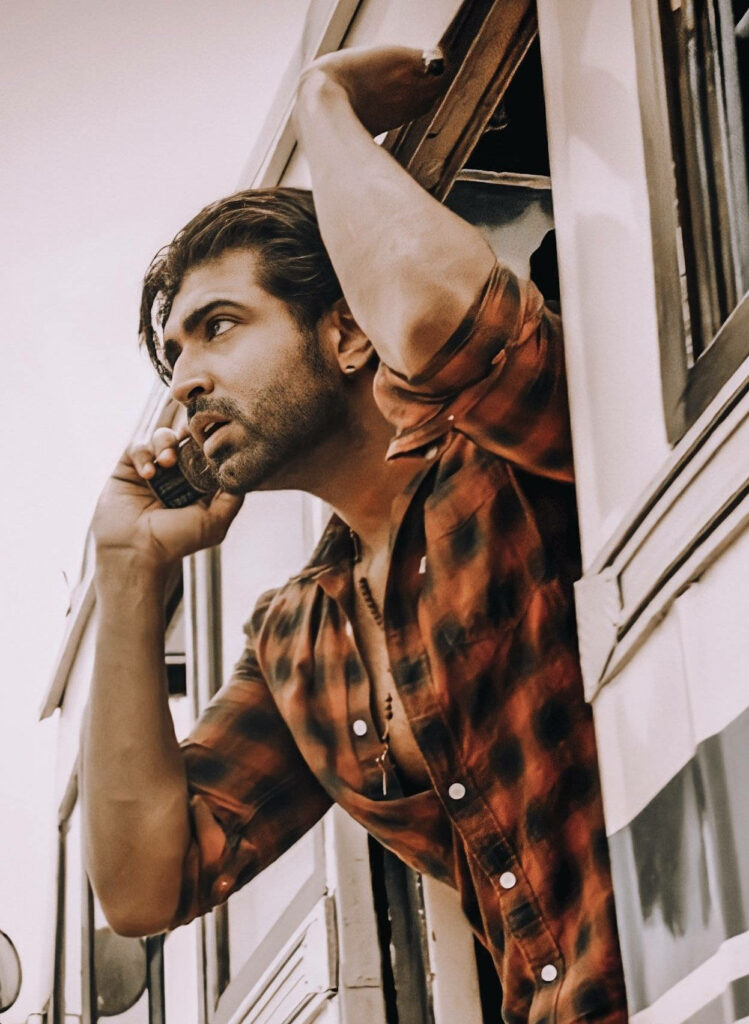 Arun Vijay in checked shirt making intense phone call leaning out bus window with wallpaper background