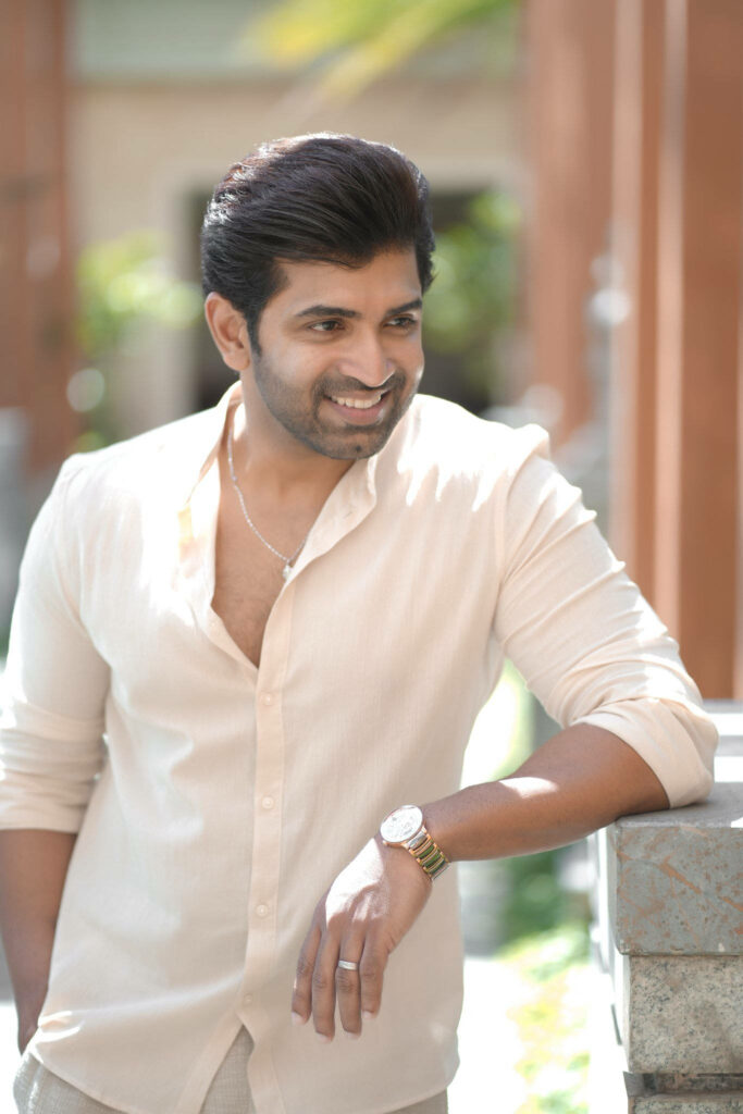 Arun Vijay in peach shirt smiling warmly leaning on stone railing bright natural wallpaper