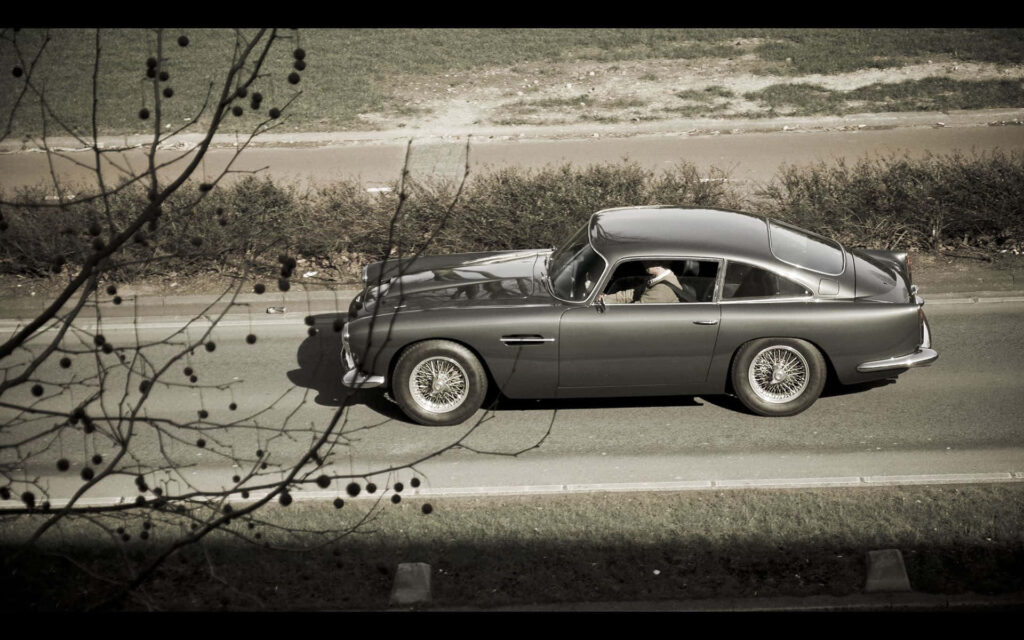 Aston Martin DB5 classic luxury car driving smoothly on a quiet road wallpaper background