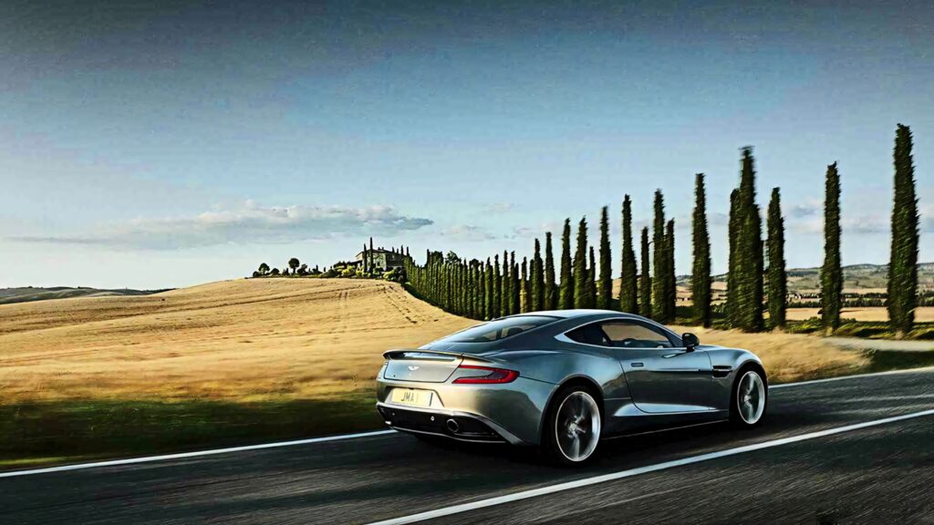 Aston Martin DB9 driving on scenic countryside road with golden fields and cypress trees Wallpaper