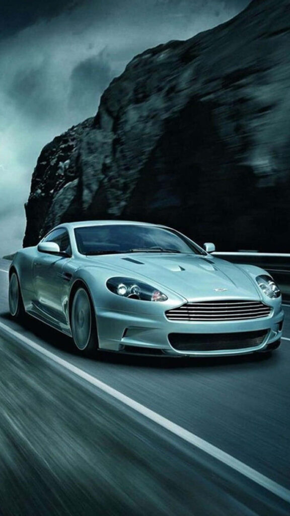 Aston Martin DB9 driving fast on a winding mountain road with dramatic cloudy sky background Wallpaper