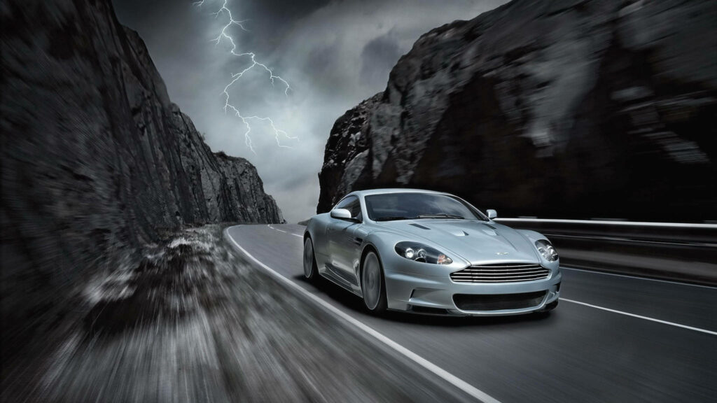 Aston Martin DBS driving on winding mountain road with stormy sky and lightning background Wallpaper