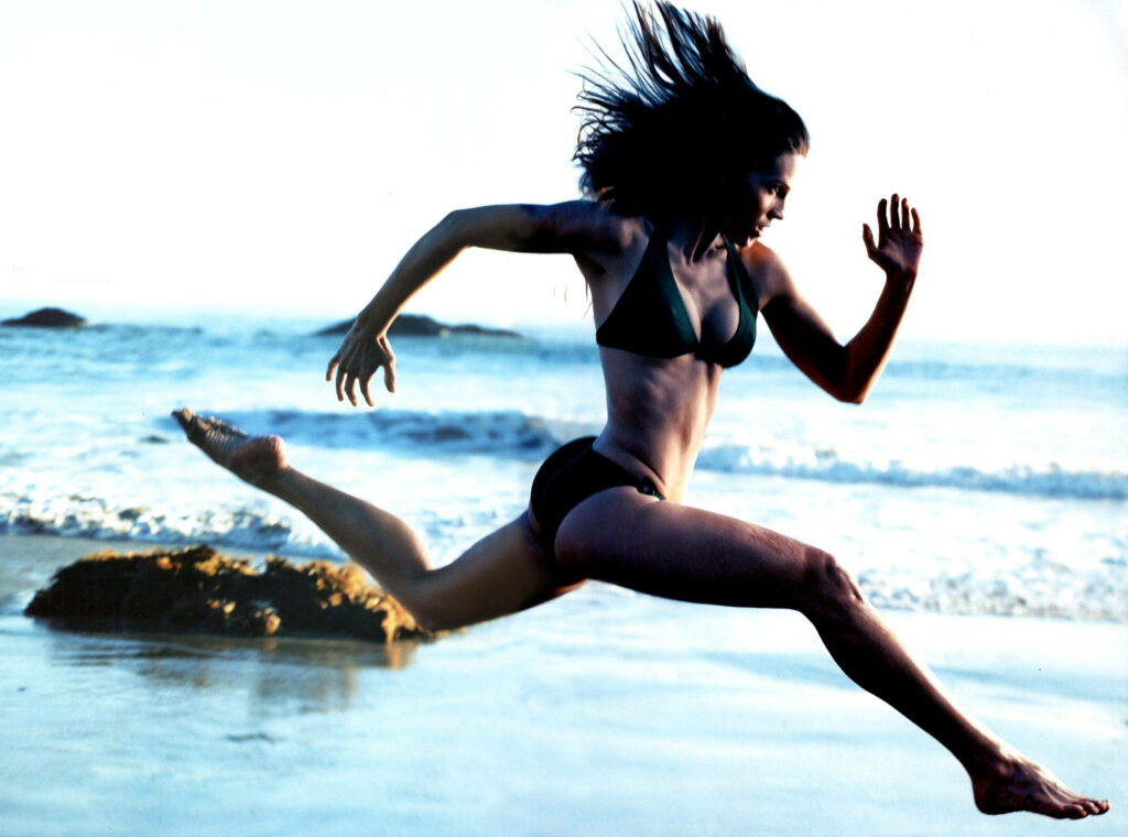 Athletic woman in black bikini running and leaping on beach with waves rocks ocean background Wallpaper