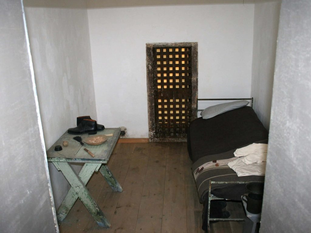 Historical prison cell wallpaper with cot wooden table barred door and stark lighting background