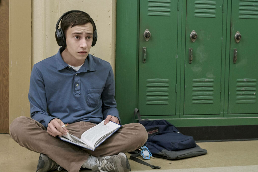Atypical lead Sam focused reading and taking notes cross-legged by green lockers with headphones Wallpaper