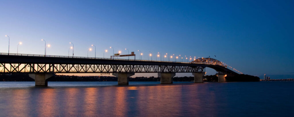 Auckland Harbour Bridge illuminated at dusk reflecting on calm waters with clear evening sky Wallpaper