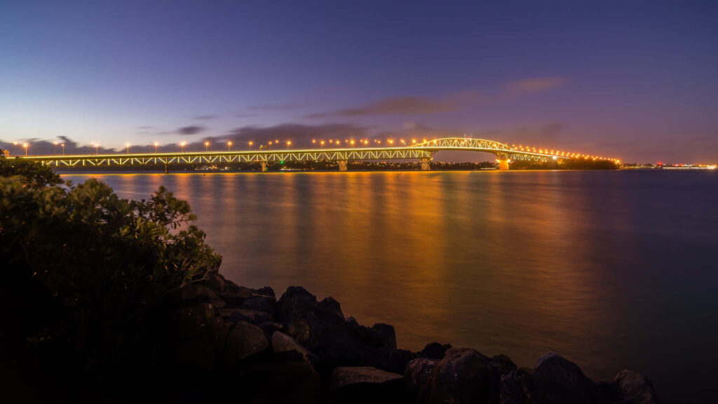 Auckland Harbour Bridge illuminated with golden lights reflecting on calm water at night Wallpaper