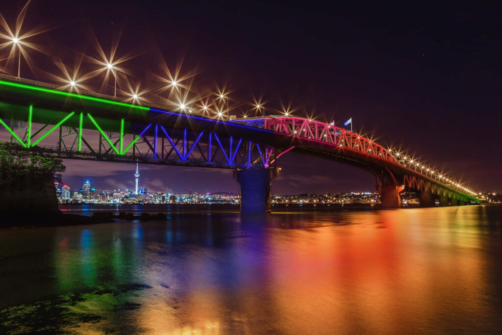 Auckland Harbour Bridge lit up at night with colorful lights reflecting on calm water and city skyline Wallpaper