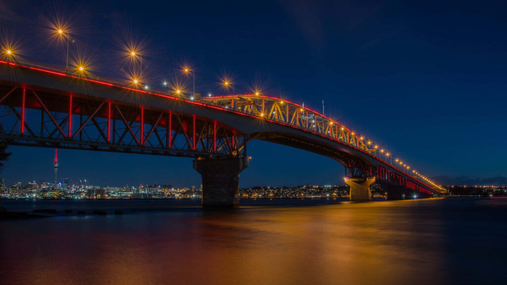 Auckland Harbour Bridge lit up at night reflecting on calm water with city skyline background Wallpaper