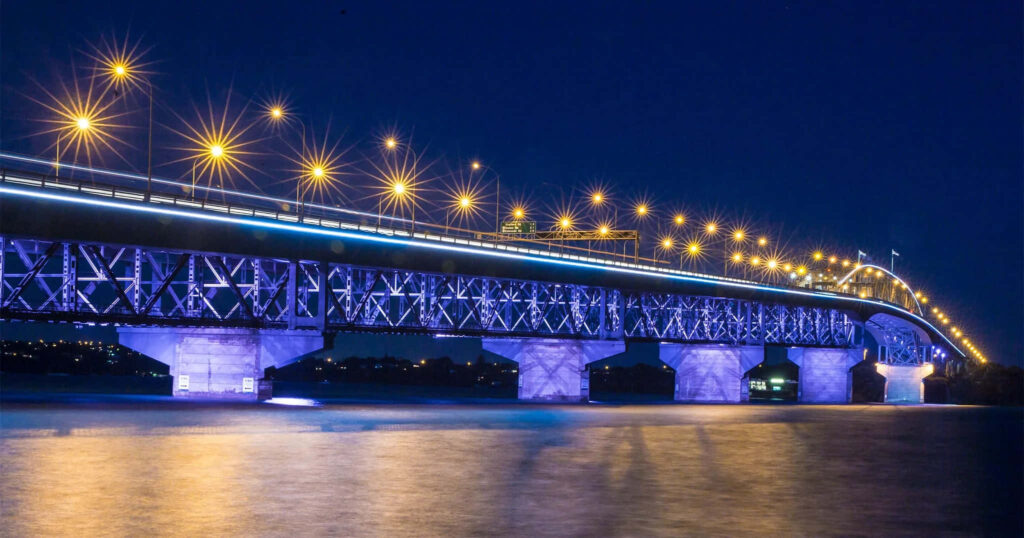 Auckland Harbour Bridge illuminated at night reflecting on calm water with deep blue sky background Wallpaper