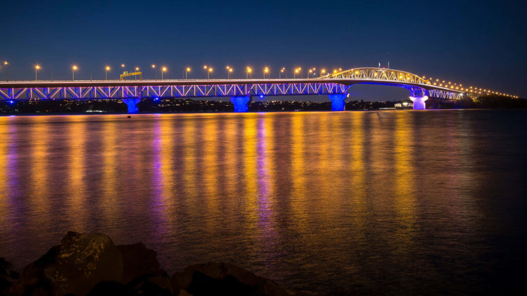 Auckland Harbour Bridge lit up at night with colorful lights reflecting on calm water wallpaper