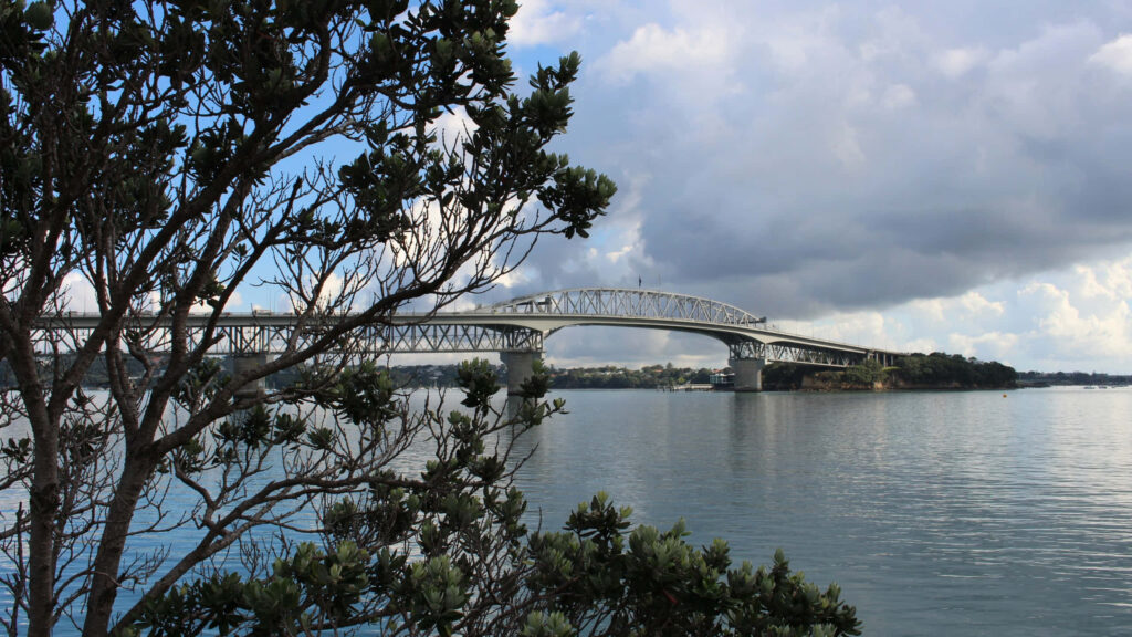 Auckland Harbour Bridge over calm water with lush greenery and dramatic sky wallpaper background