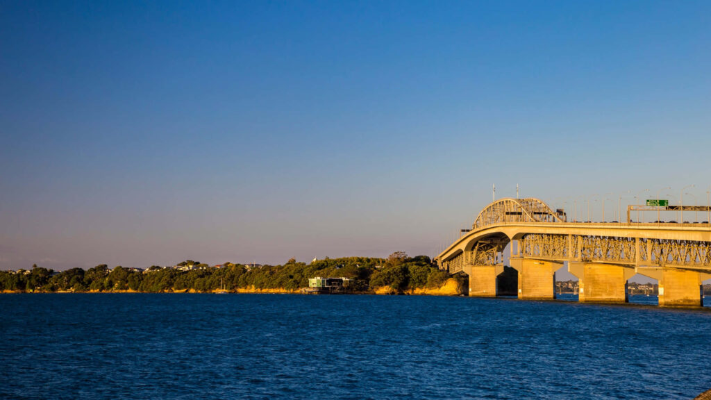 Auckland Harbour Bridge at sunset over calm blue waters with golden light and clear sky background Wallpaper