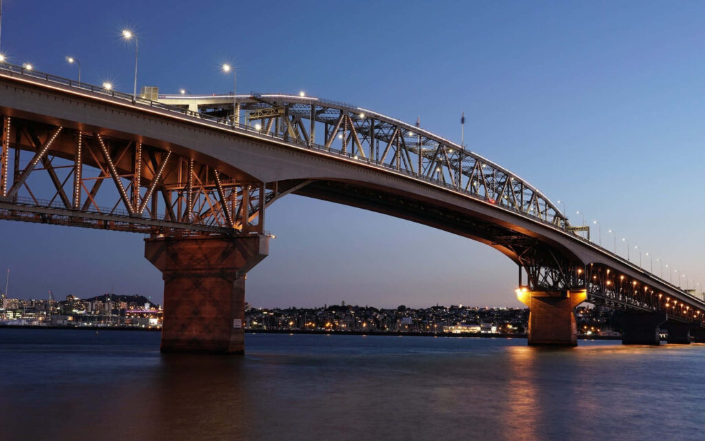 Auckland Harbour Bridge glowing at twilight reflecting city lights on calm water serene scene Wallpaper