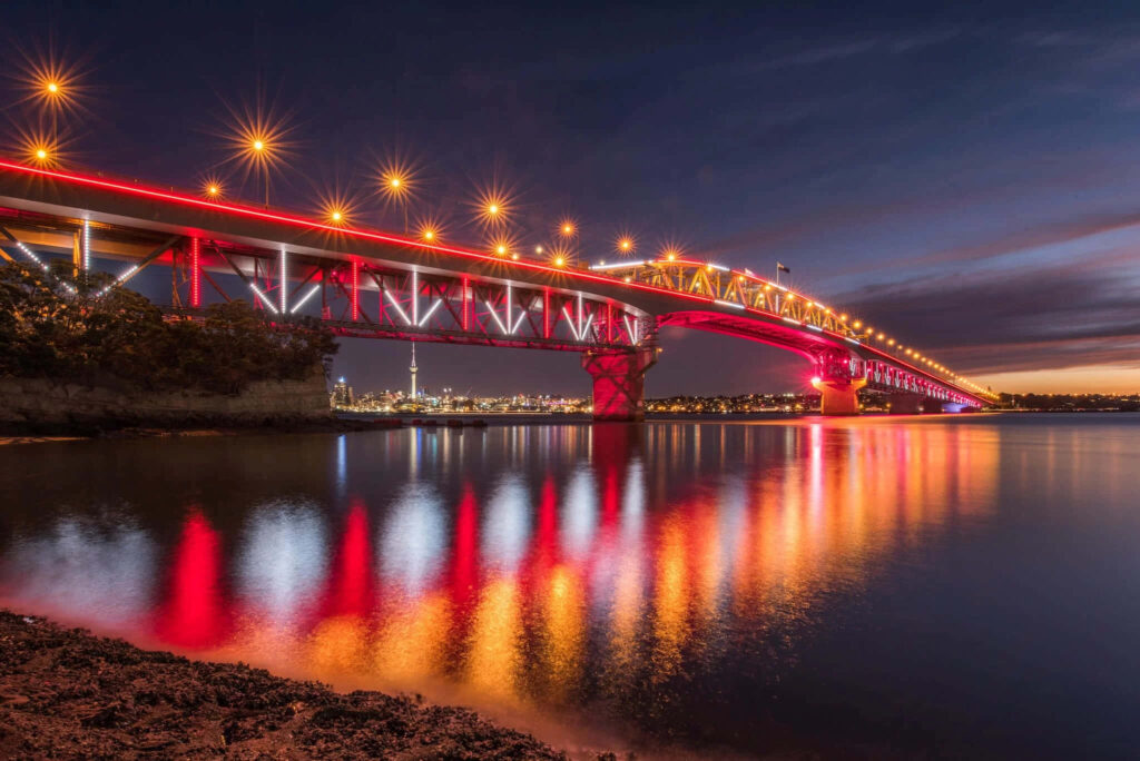 Auckland Harbour Bridge illuminated with vibrant lights at twilight reflecting on calm waters Wallpaper