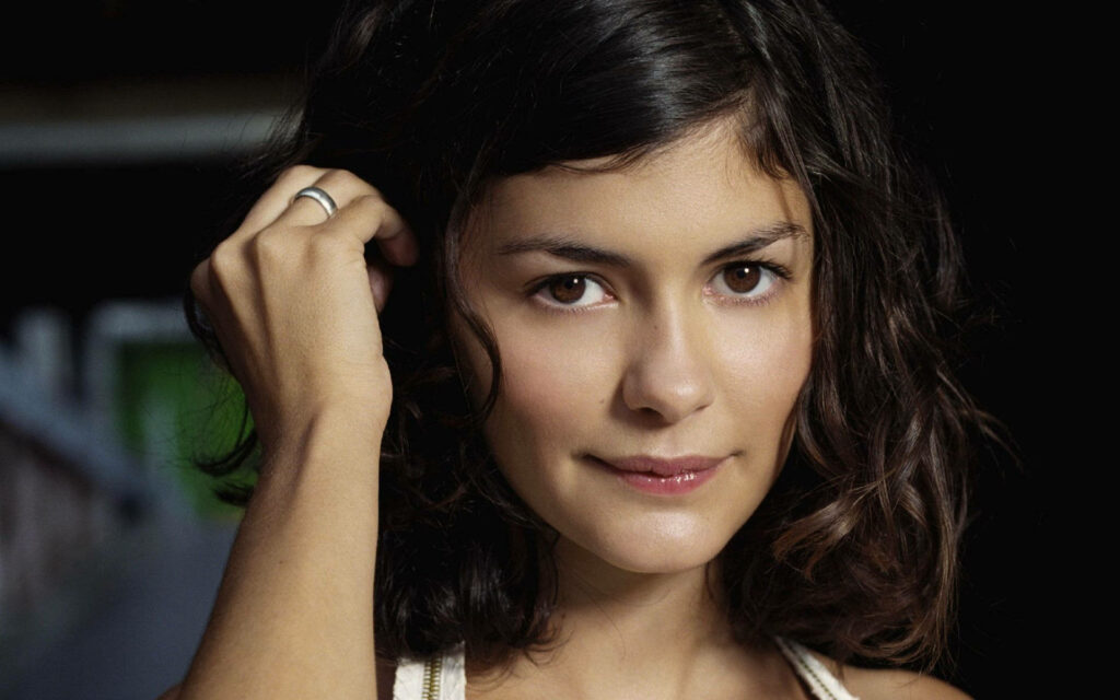 Audrey Tautou gently pushing hair back with soft inviting expression dark wallpaper background